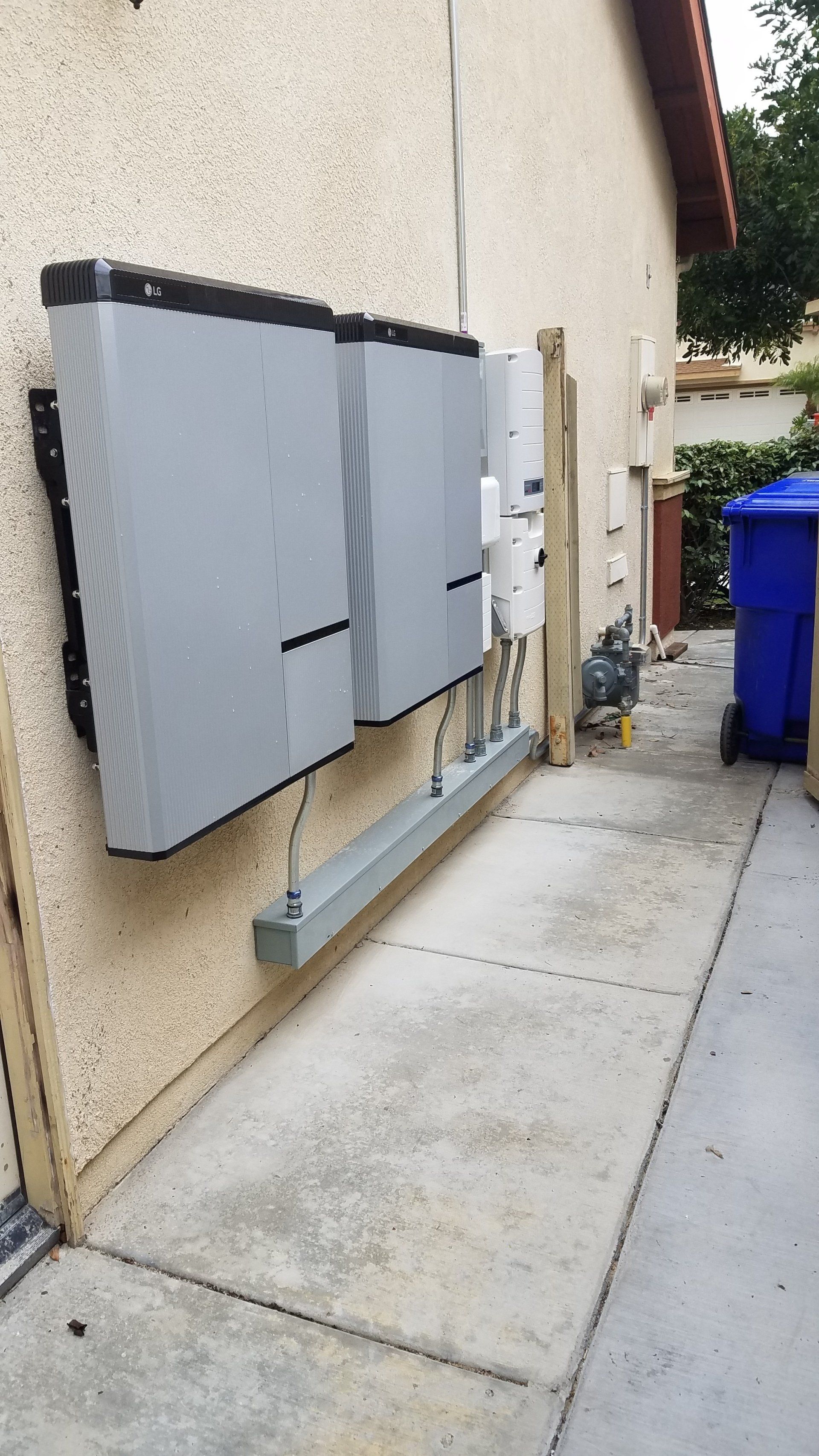 Solar power equipment mounted on a beige building exterior, next to a trash bin on a concrete walkway.