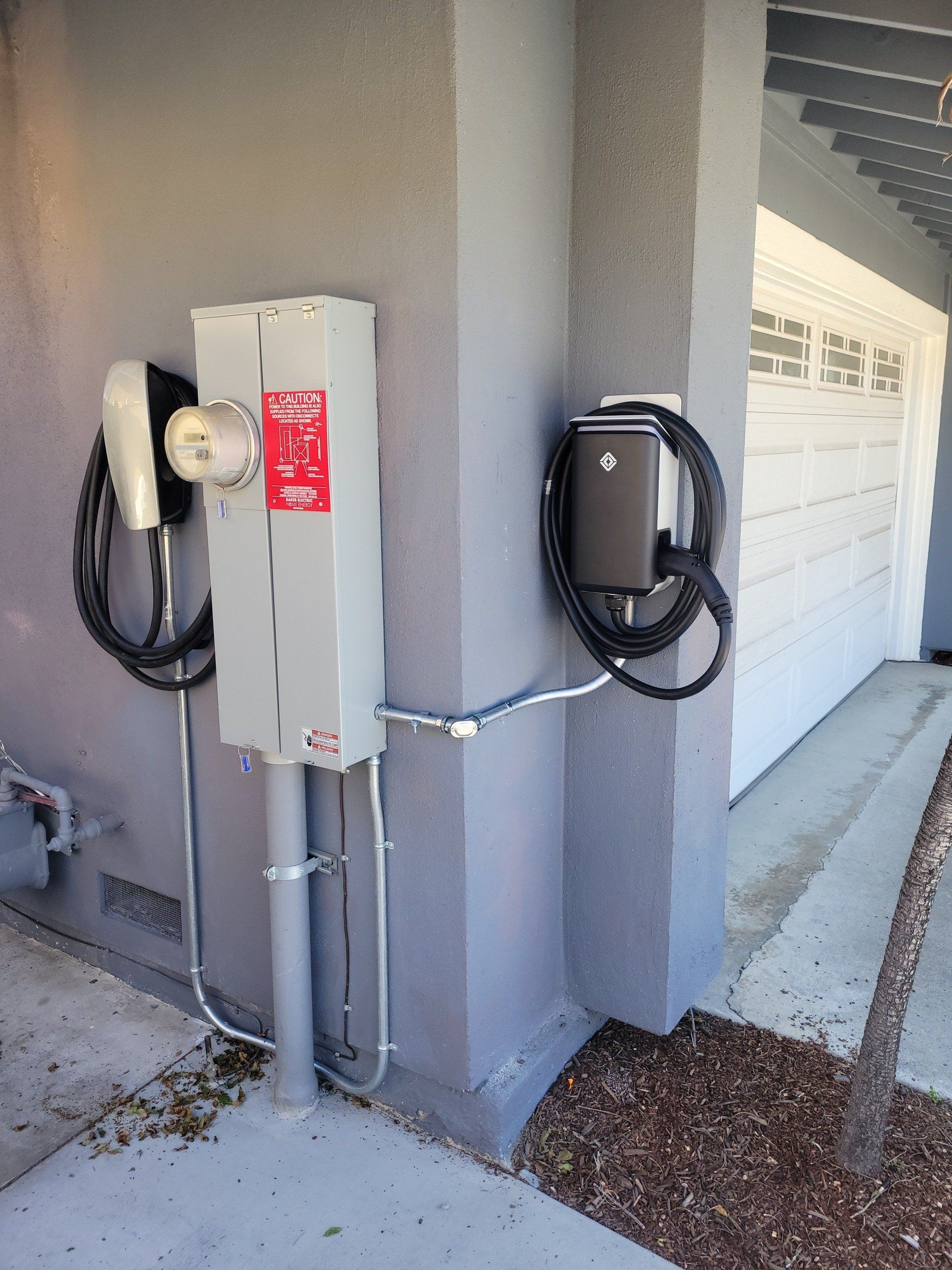 Gray electrical boxes and EV chargers mounted on a gray stucco exterior near a garage.
