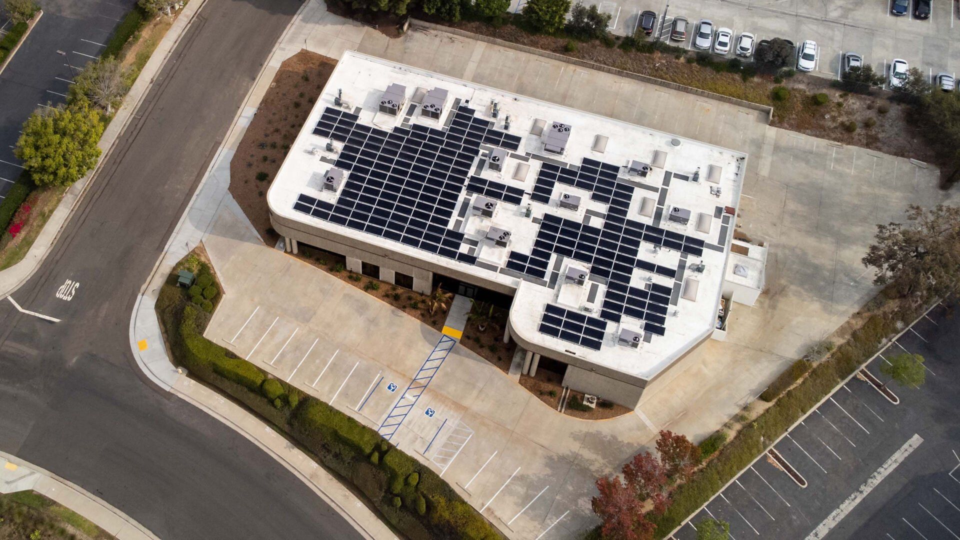 Building with solar panels on the roof, surrounded by a parking lot, trees, and a road.