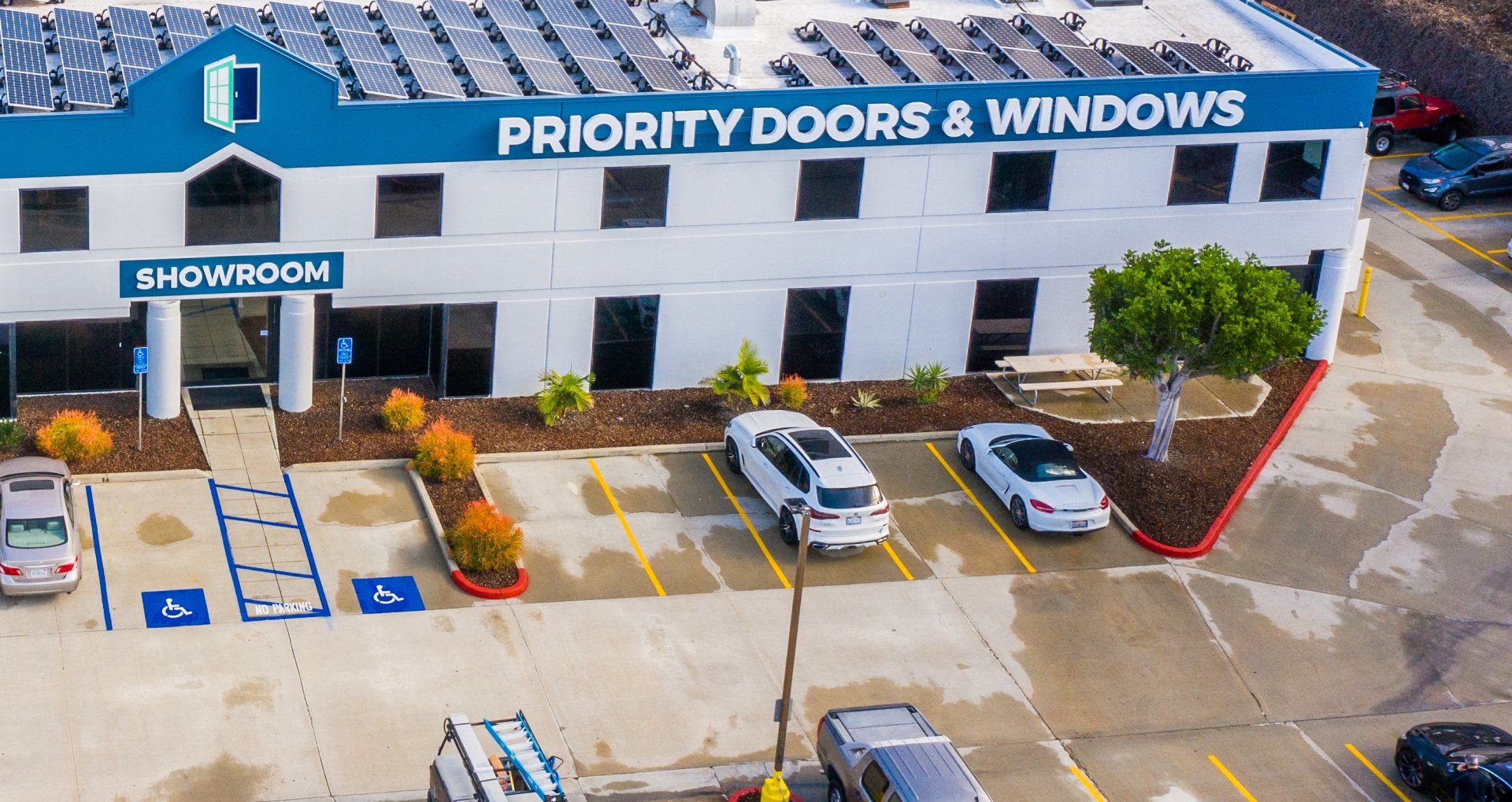 Exterior of Priority Doors & Windows showroom with parked cars, blue and white building, and solar panels on the roof.