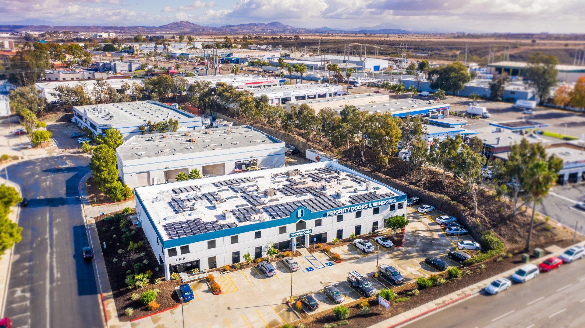 Aerial view of an industrial park, with buildings, trees, and cars. Solar panels on a large white building.