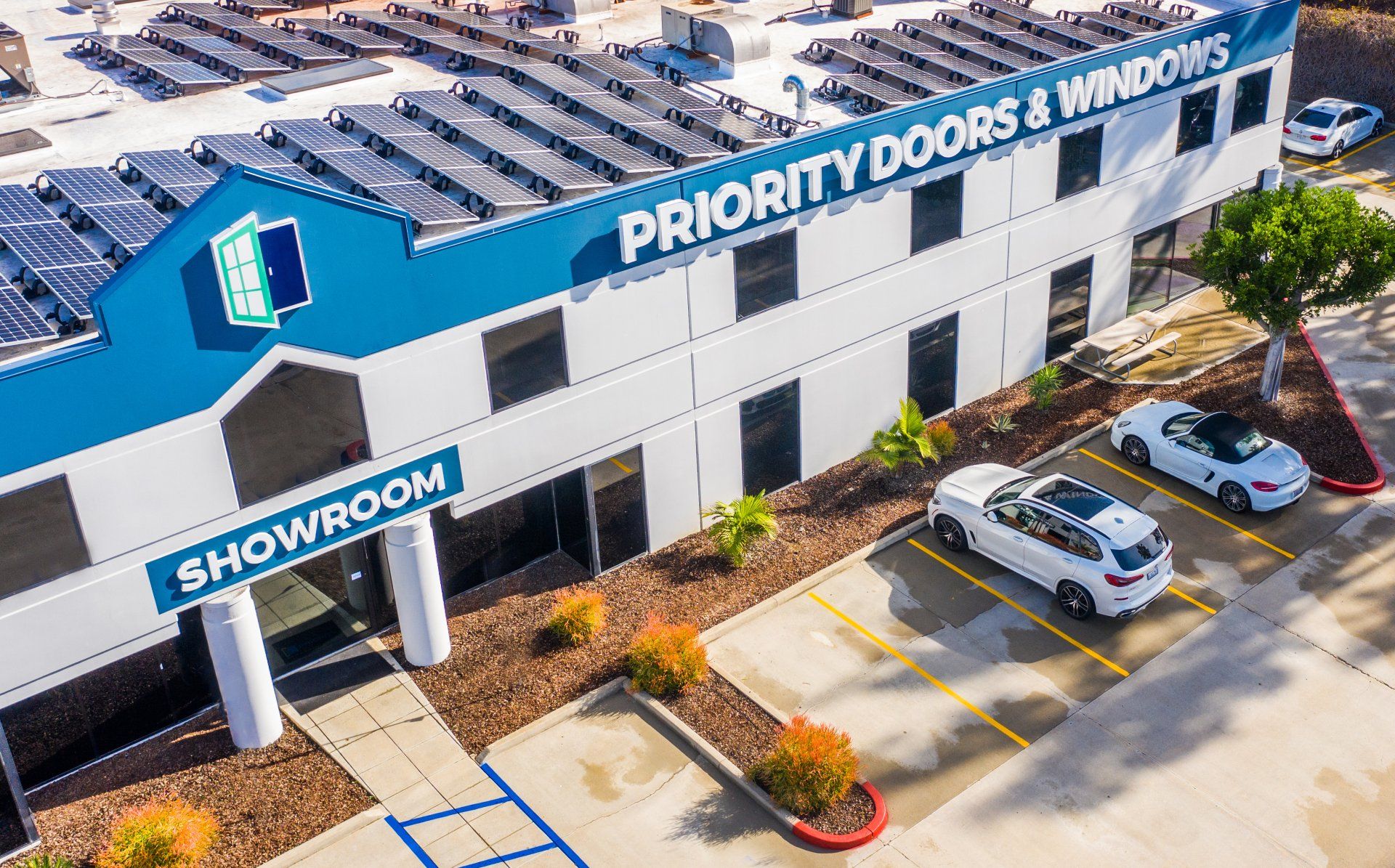 Exterior view of Priority Doors & Windows showroom, blue and white building with solar panels, cars parked outside.