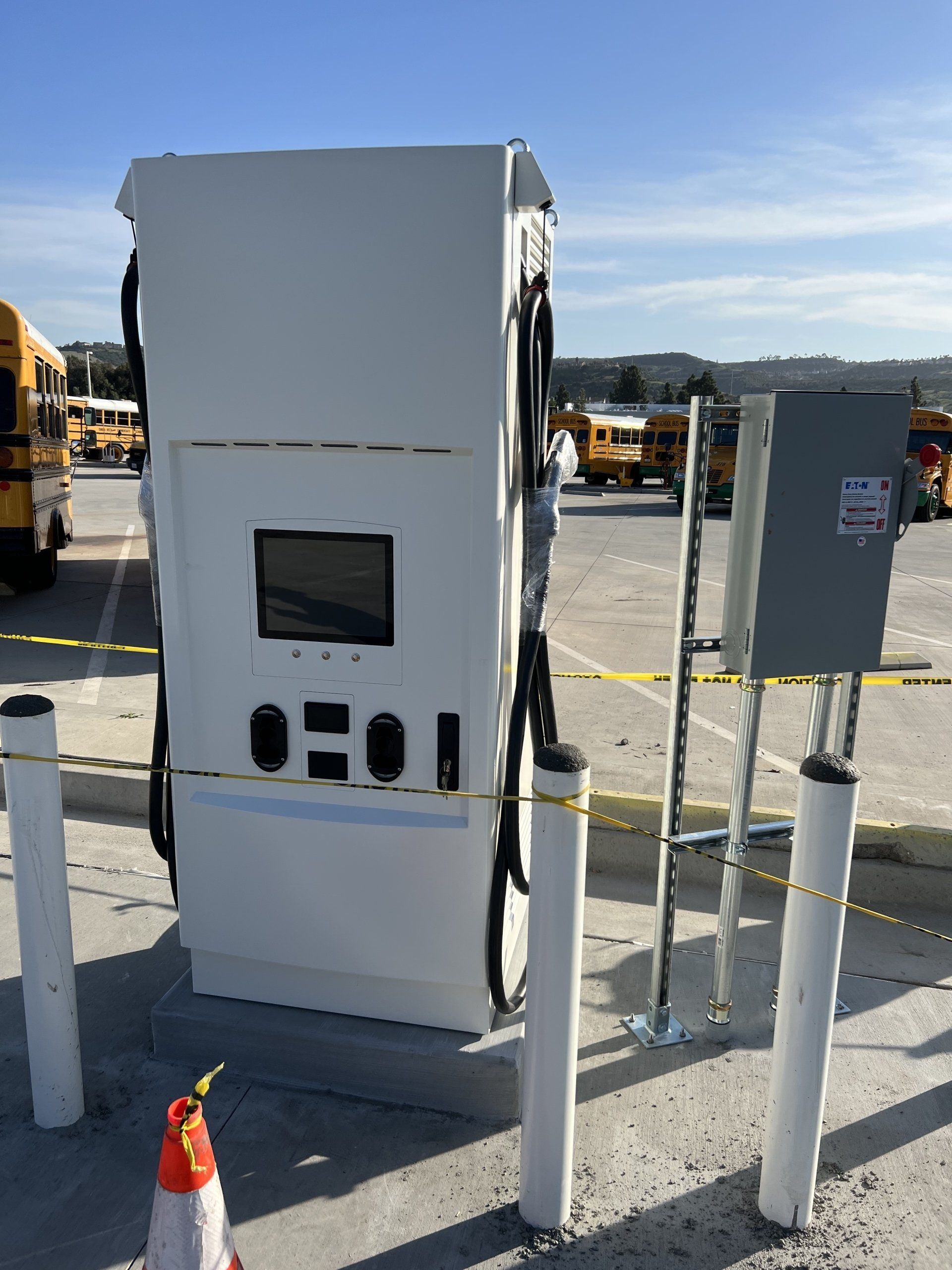 White electric bus charging station with multiple cables, in a parking lot with school buses.