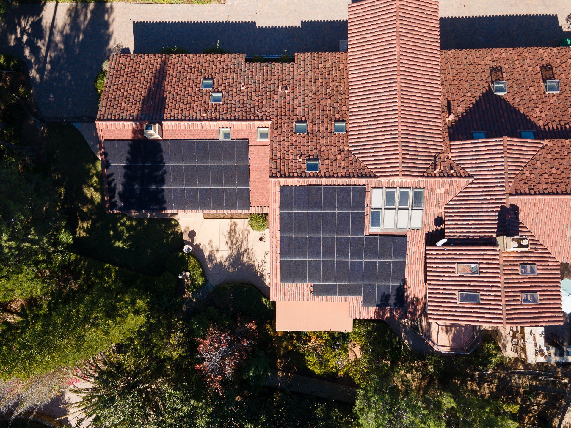 Overhead view of a house with solar panels on a red tile roof, surrounded by trees.