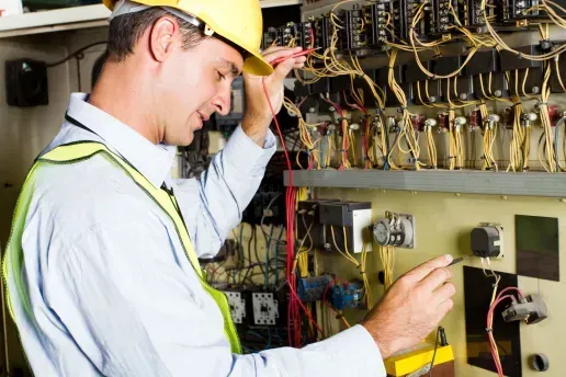 Electrician in yellow helmet and vest tests wiring on a control panel.