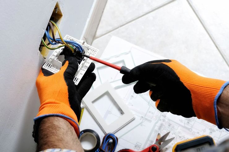 Electrician wearing orange gloves works on electrical wiring inside a wall, using a screwdriver.