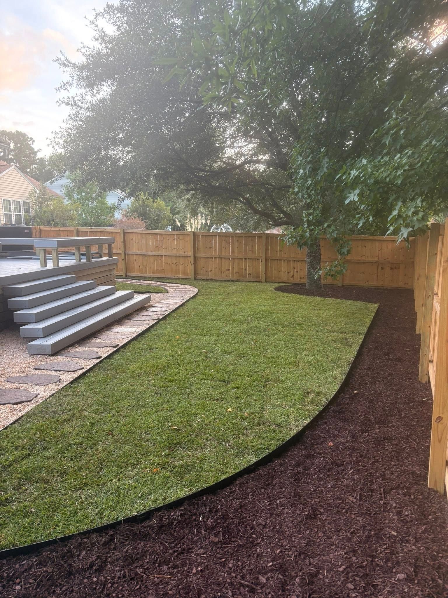 Backyard with curved lawn border, stone steps, wooden fence, and a large tree under overcast light
