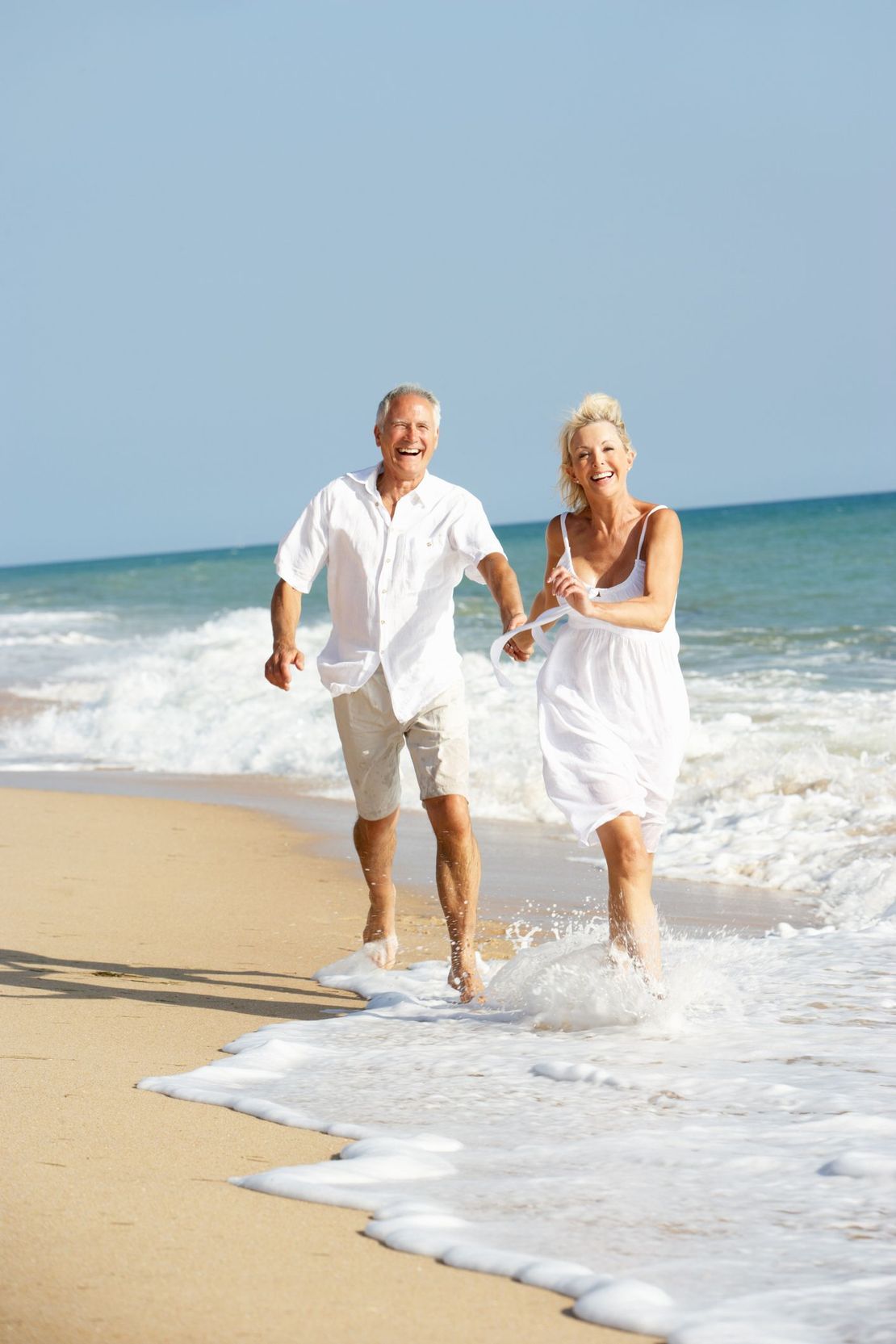 Two people in white clothes run barefoot along a sunny beach with waves behind them.