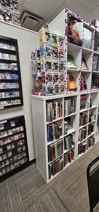 A retail store displaying DVDs, Funko Pops, and collectible trading cards on shelves.