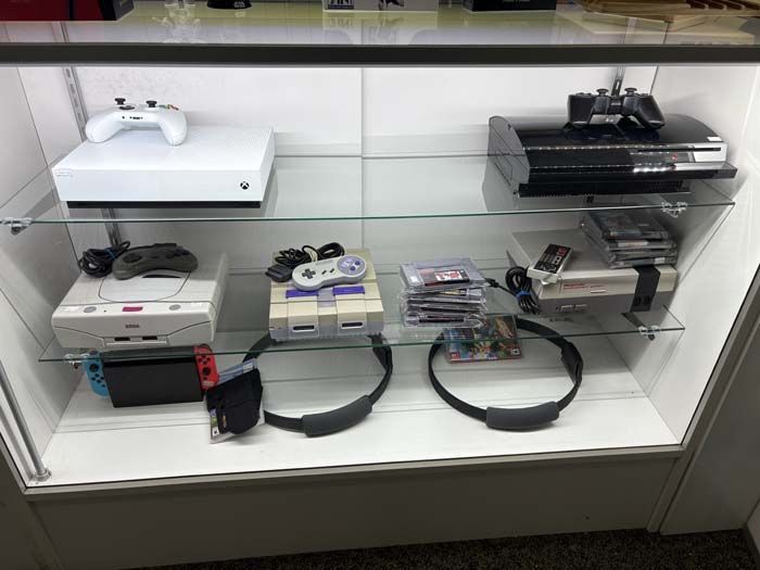 Video game consoles and accessories displayed in a glass case: Xbox, PlayStation, Nintendo Switch and older consoles.
