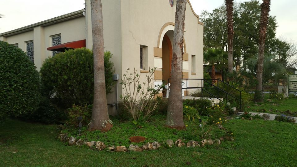 A light-colored stucco building with an arched entrance, framed by two tall palm trees and a garden bed in the foreground.
