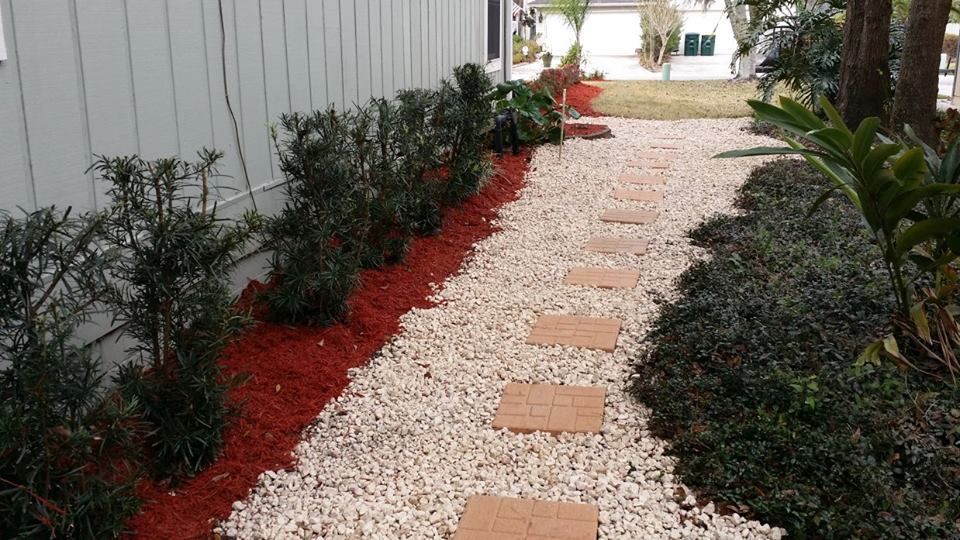 A stone path with square stepping stones lined by red mulch and shrubs next to a gray building wall.
