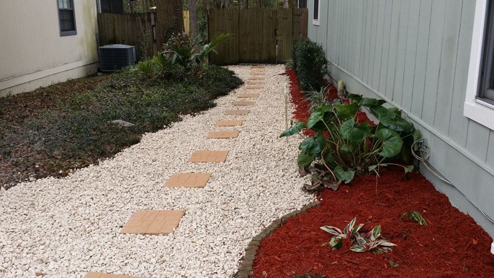 A gravel pathway with stepping stones leads between a house and garden beds with red mulch and green plants.