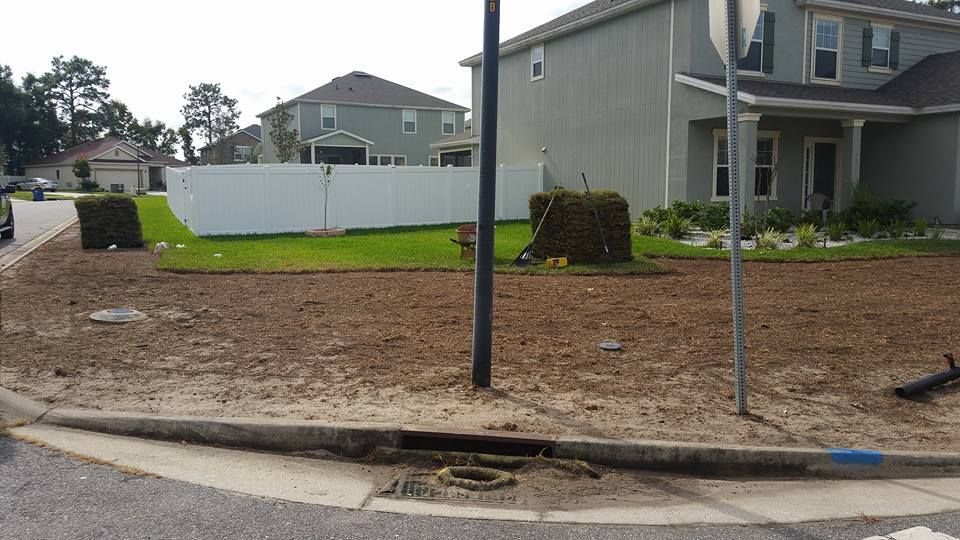 A street-level view of a residential corner lot with a dirt yard, green lawn, white vinyl fence, and a storm drain.