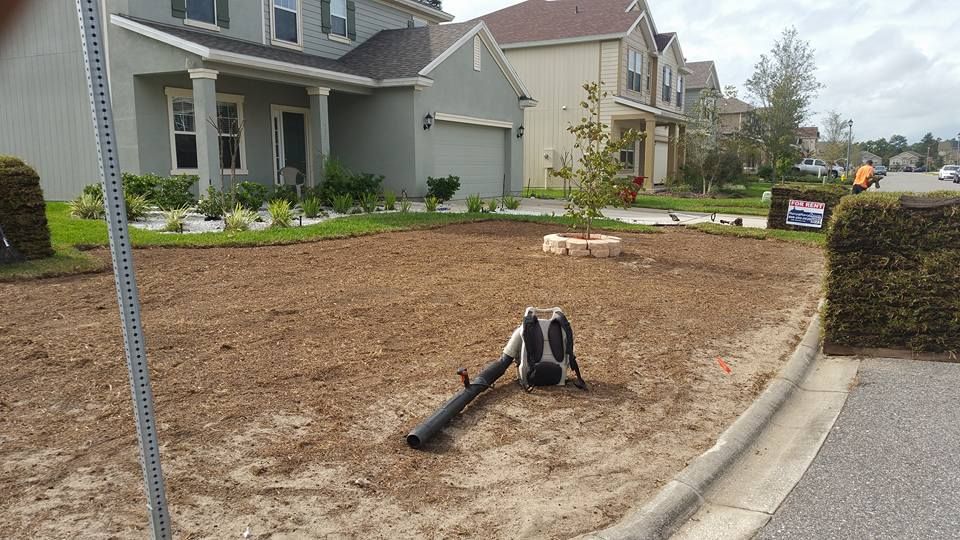 A backpack leaf blower rests on a bare, dirt-covered front yard of a suburban house.