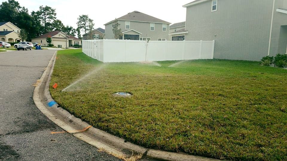 A sprinkler waters a residential lawn along a street curb in a suburban neighborhood with houses and a white fence.