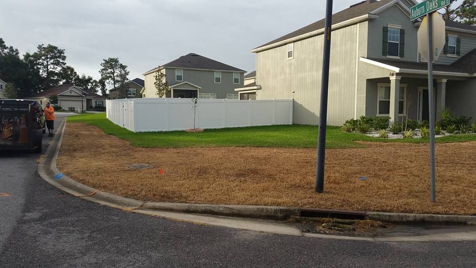 A residential street corner featuring a white vinyl fence, green grass, and brown mulch landscaping near suburban homes.