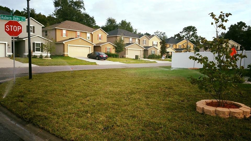 A sprinkler waters a residential lawn next to a street sign and a stop sign in a suburban neighborhood.