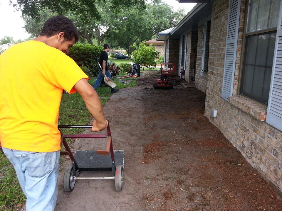 Two people operate landscaping equipment on a residential lawn next to a brick house.