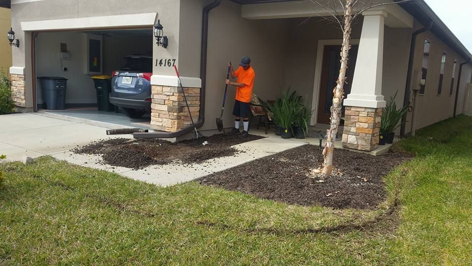 A person in an orange shirt shovels dark mulch into a garden bed in front of a house with a garage and stone pillars.