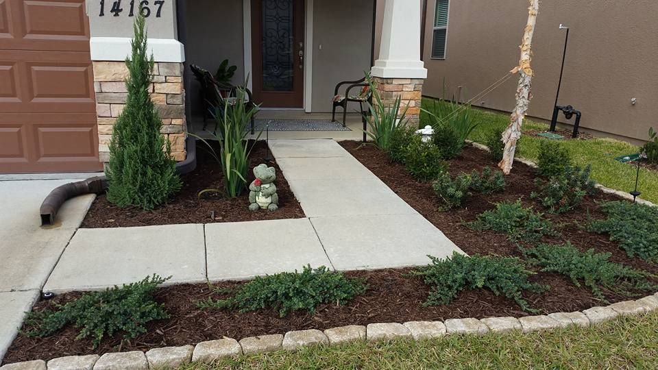 A concrete walkway leads to a front door, flanked by mulched garden beds with shrubs and decorative stone edging.