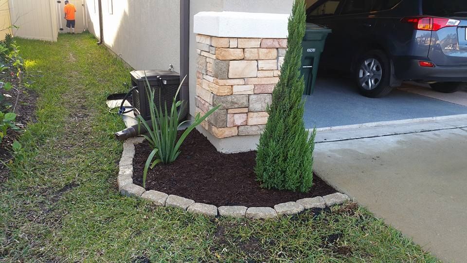 A garden bed with a small conifer and grass in front of a stone-clad house pillar near a driveway.
