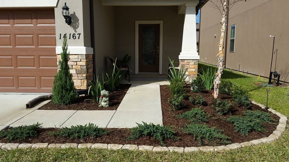 A suburban home entryway featuring a concrete walkway, garden beds with shrubs, mulch, and stone edging.