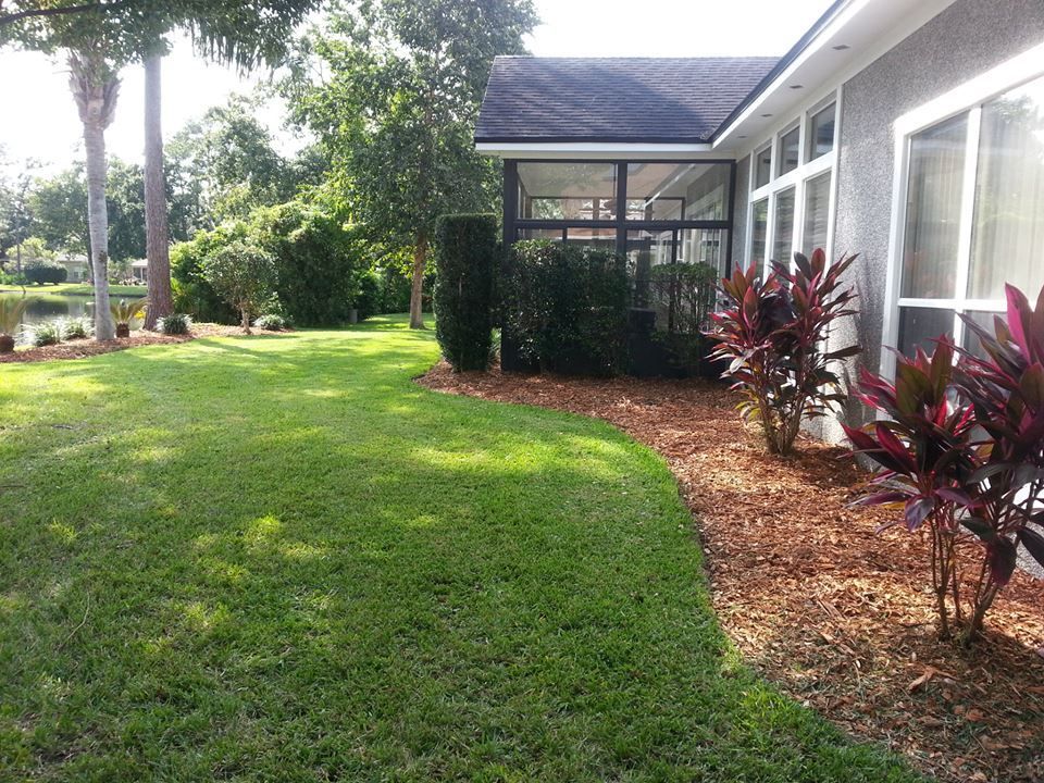 A side view of a house with a screened porch, a mulch garden bed with red plants, and a manicured lawn by a lake.