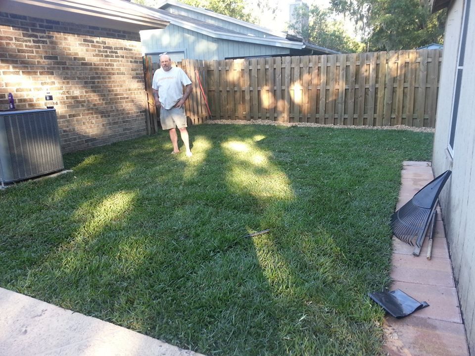 A man standing in a residential backyard with green grass, a brick wall, and a wooden privacy fence.