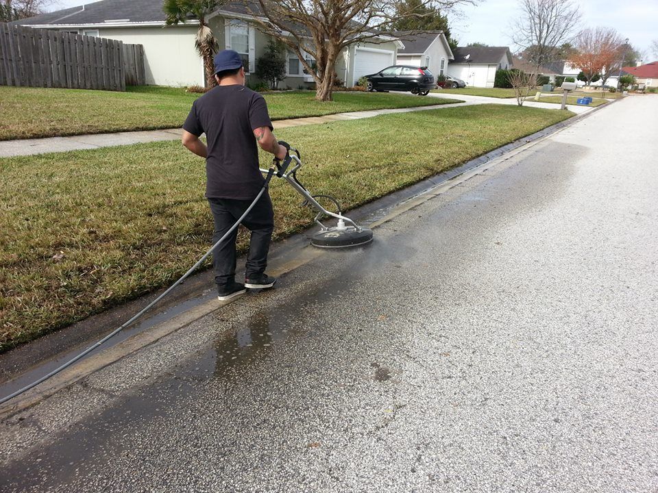 A person uses a pressure washer surface cleaner on a concrete curb along a suburban street.