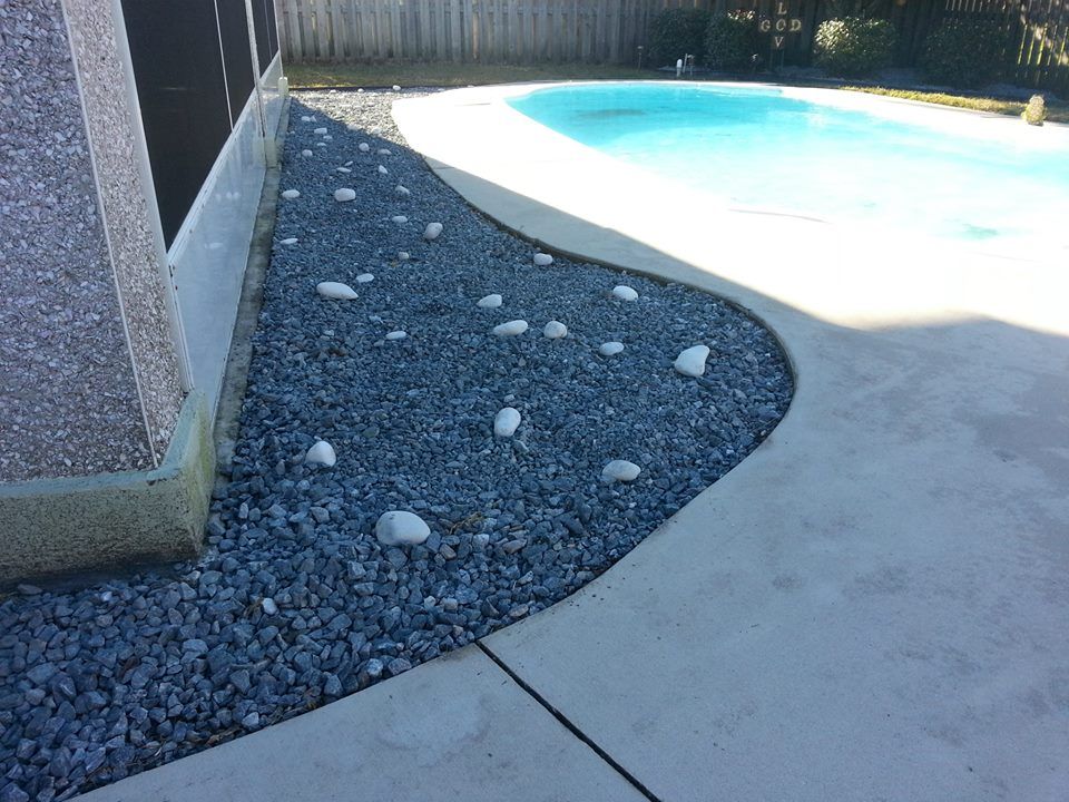 A curved bed of grey gravel with scattered white decorative stones next to a swimming pool and a house wall.