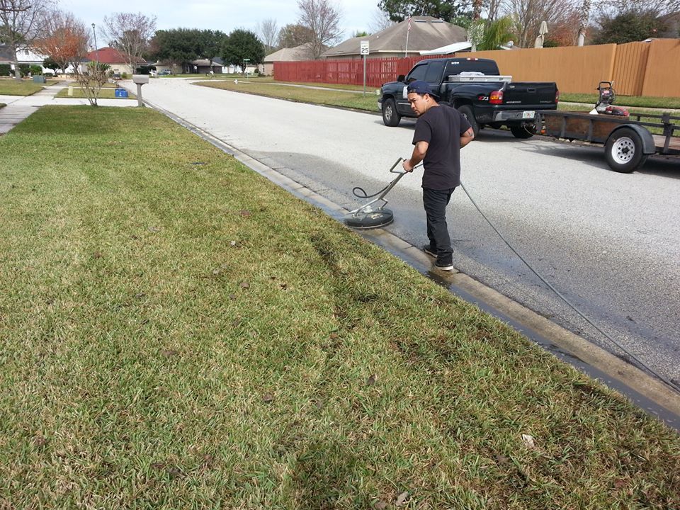 A person uses a pressure washer to clean a concrete curb along a residential street next to a grassy lawn.