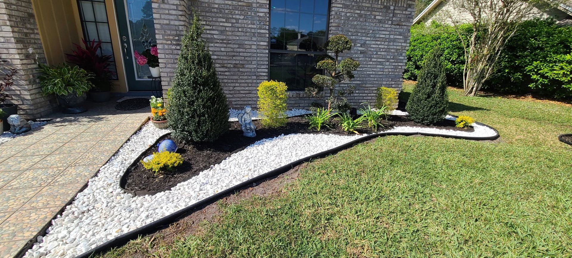 Curved white gravel bed with shrubs and black edging beside a brick house and green lawn