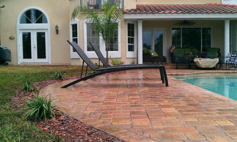 Two lounge chairs sit on a brick patio next to a pool outside a house.