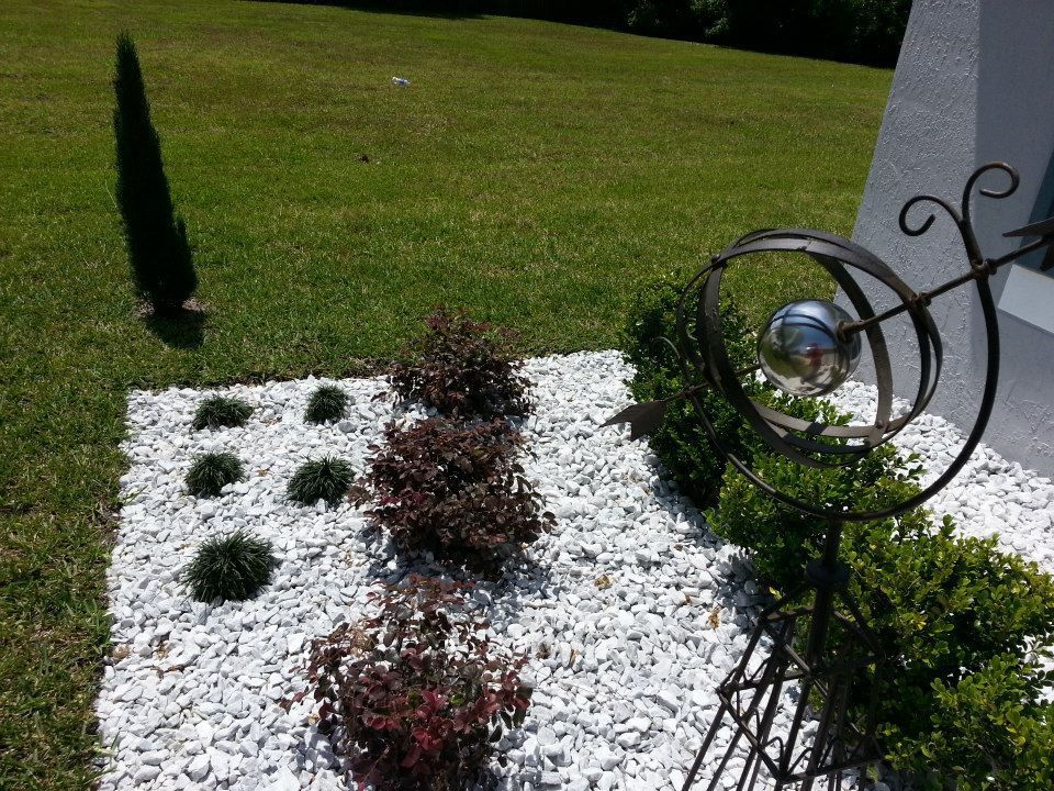 A white rock garden bed features small dark-leaf shrubs and a decorative metal sculpture, beside a green lawn.