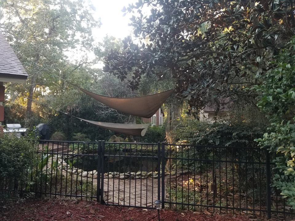 A backyard area featuring a pond and stone border, framed by trees with tan shade sails hanging above a black metal fence.