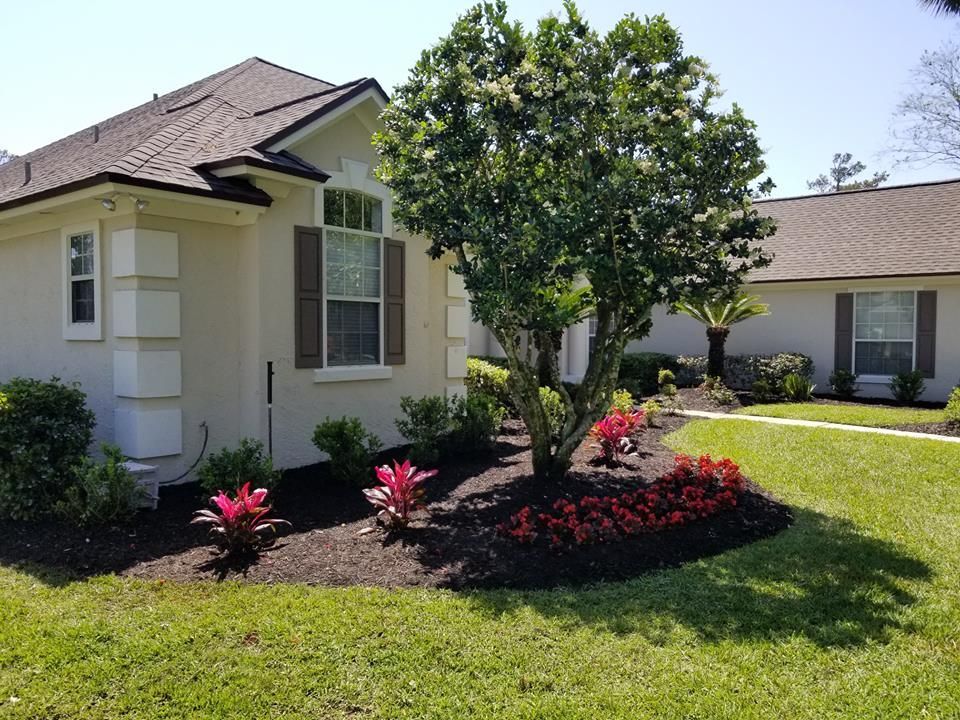 A light-colored house with a dark roof and brown shutters, fronted by a garden with a central tree and red plants.