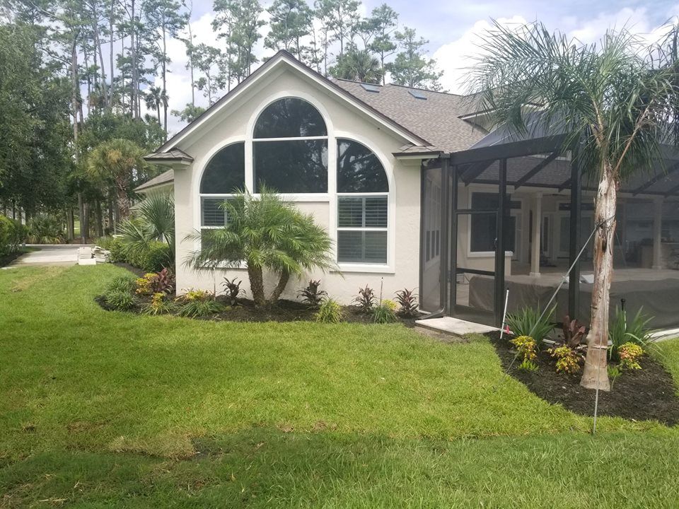 A single-story home with a large arched window, a screened-in lanai, and landscaped garden beds on a sunny day.