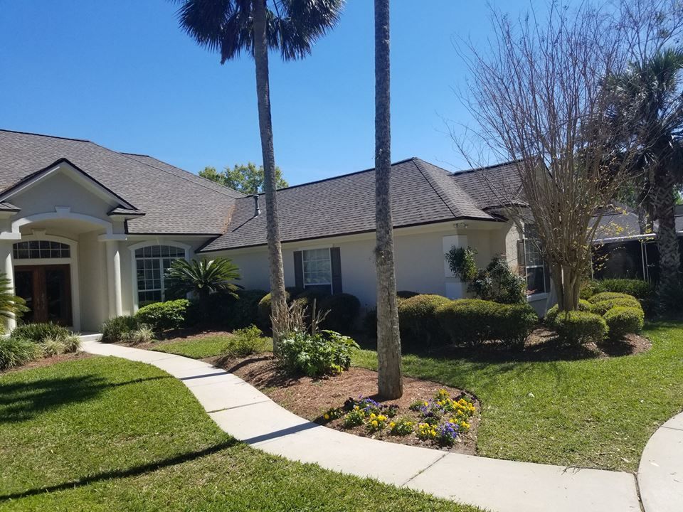 A beige house with a grey shingled roof, palm trees, and a curved concrete walkway leading to the front entrance.