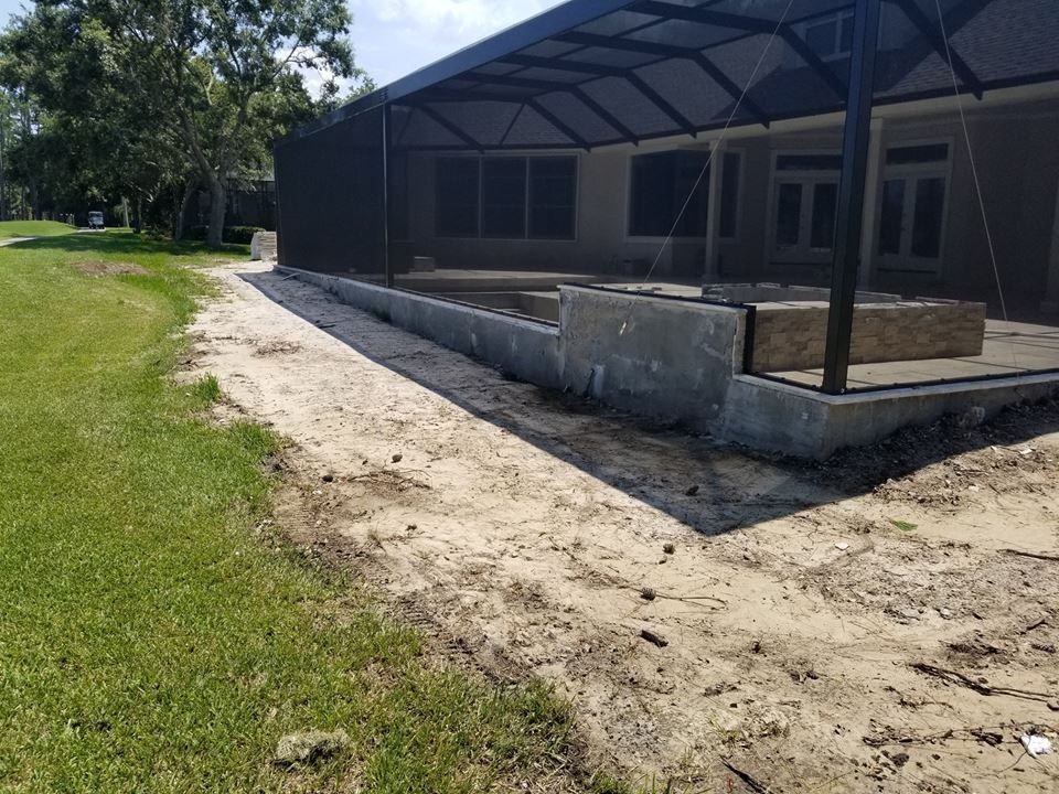 A screened-in patio with a concrete retaining wall under construction, bordered by a dirt path and a grassy lawn.