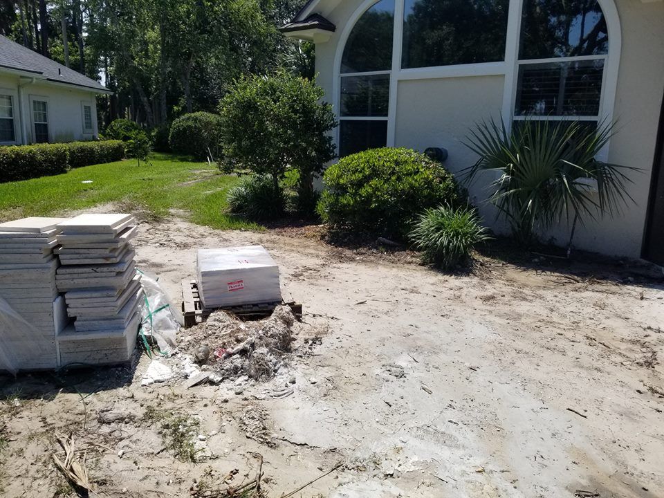 Two stacks of paving tiles sit on a sandy ground beside a house with arched windows and bushes.