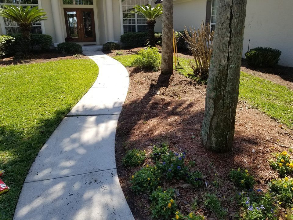 A winding concrete path leads to a home entrance, flanked by a green lawn and a garden bed with trees, mulch, and flowers.