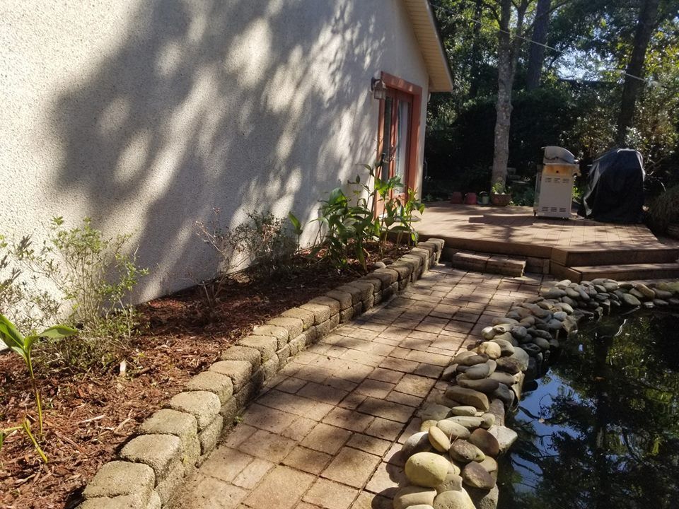 A brick path runs between a garden bed with stone edging and a water feature next to a house with a wooden deck.