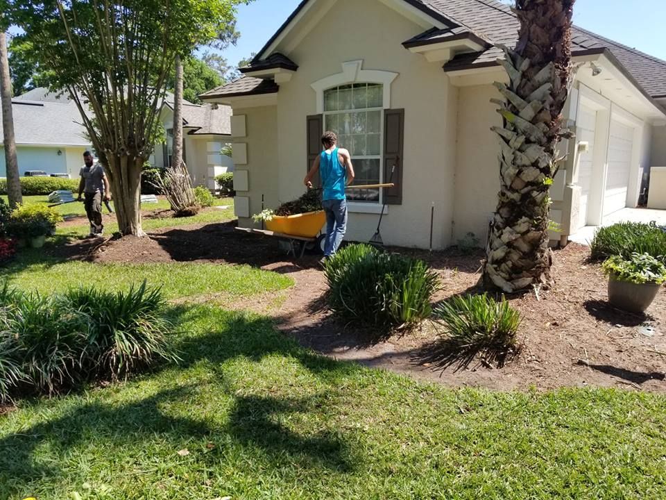 Two workers spread mulch in the front garden beds of a single-story house on a sunny day.