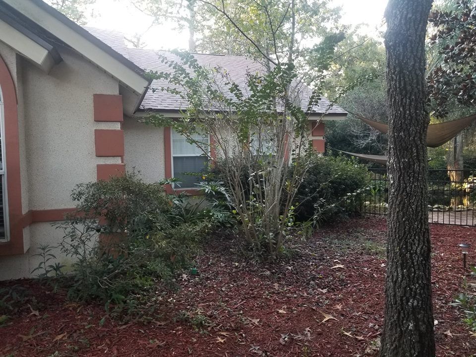 Exterior view of a stucco house with rust-colored accents, surrounded by mature landscaping and a mulch-covered yard.
