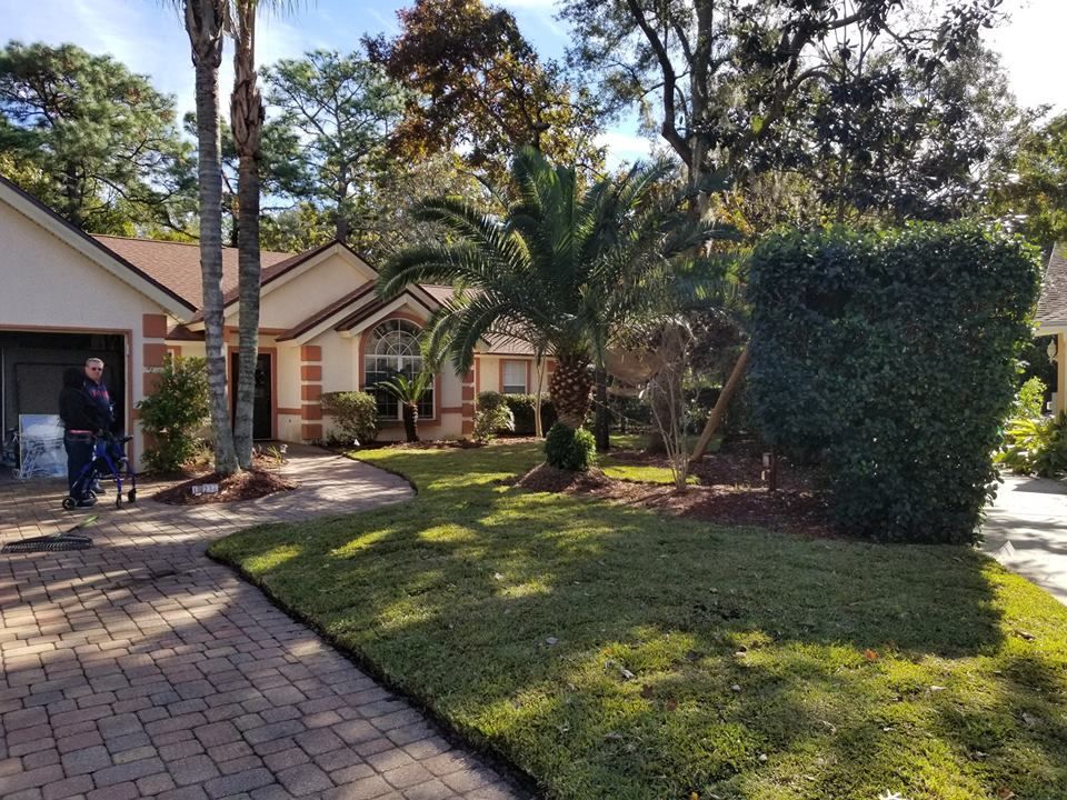 A person in dark clothing stands near a garage door in front of a house with a brick driveway and landscaped lawn.