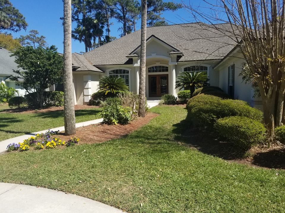 A one-story suburban home with a gray roof, beige siding, a columned entrance, and a landscaped front yard with trees.