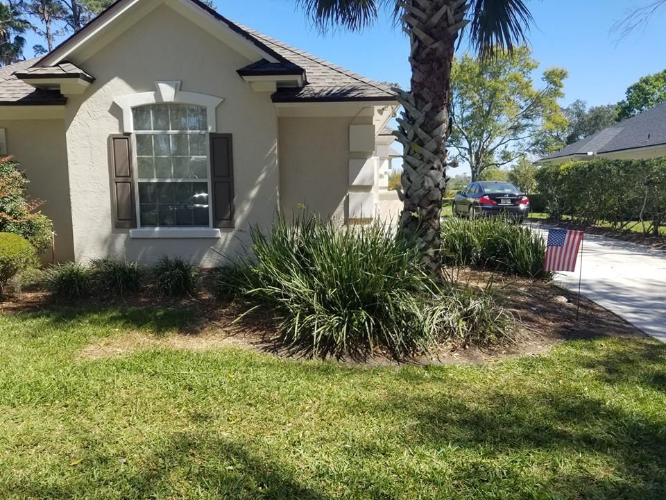 A beige house with dark shutters and a large palm tree in the front garden, with a small American flag by the driveway.