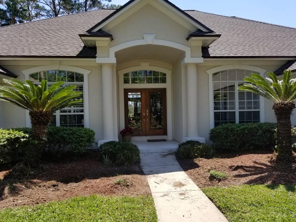 A beige, single-story house with a wooden double front door, white columns, and palm trees flanking the entrance.