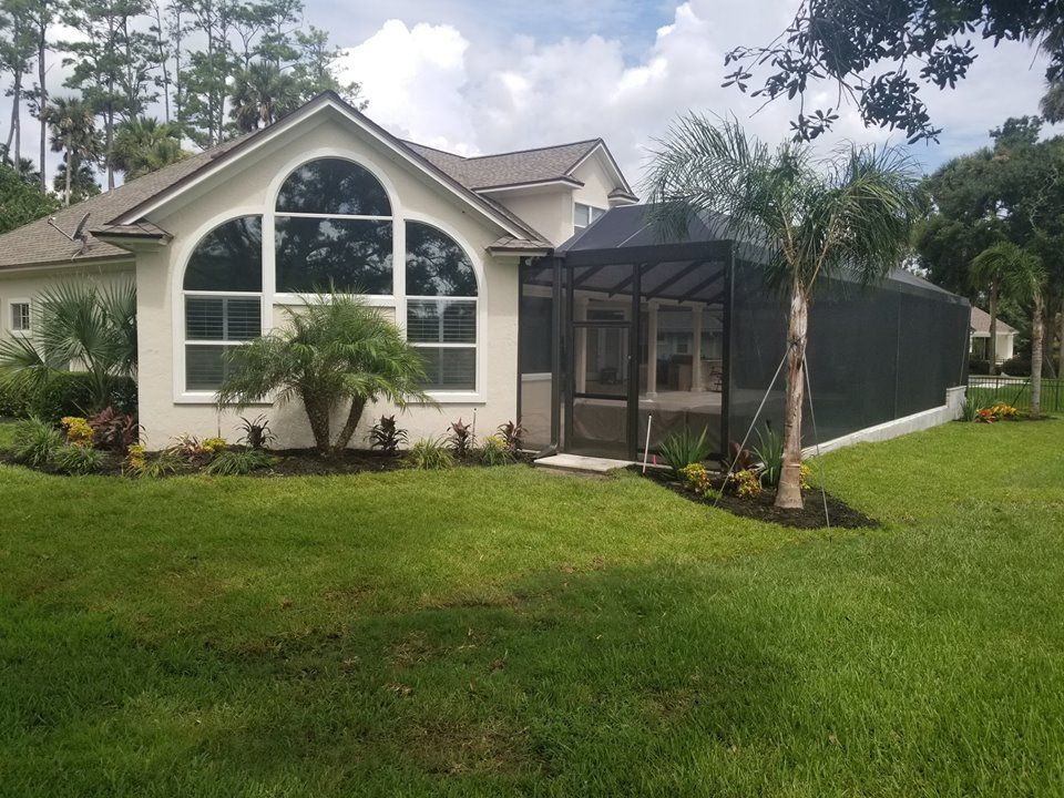 A beige house with a large arched window and a screened-in lanai surrounded by a green lawn and tropical landscaping.